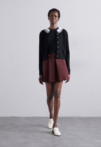 Juodas megztinis su balta nėriniuota apykakle, užsidėtas ant juodo marškinėlio. Bordo renkamas sijonas. Baltos loafer bateliai. Modelis stovi prieš neutralų foną.