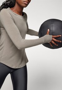 Bledě šedý sportovní top s dlouhým rukávem a zaobleným lemem, kombinovaný s černými legínami. Drží texturovanou černou medicinbal.