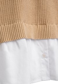 Beige gestrickter Pullover über einem weißen, geknöpften Baumwollhemd mit sichtbarer Rippstrickstruktur und Stoffdetails.