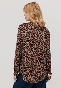 Kobieta z włosami do ramion w kolorze blond, ubrana w brązową i czarną koszulę w panterkę z długim rękawem oraz ciemnoniebieskie spodnie, stoi bokiem.