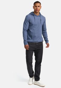 Helles blaues Kapuzensweatshirt mit gerippter Textur, langen Ärmeln und verstellbarem Kordelzug. Kombiniert mit dunklen Jeans und weißen Sneakers.