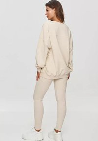 Be�żowy oversize'owy sweter z obniżonymi ramionami, prążkowanymi mankietami i dołem, w połączeniu z dopasowanymi legginsami i białymi sneakersami.