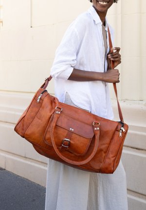 Person trägt weißes Hemd und Hose und trägt große braune Ledertasche mit Vordertasche und Schulterriemen.
