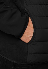 Musta takki, jossa on yhdistelmä sileää kangasta ja tikkikuvioituja osia. Taskussa on pyöreä avaus, jossa on teksturoitu yksityiskohta.