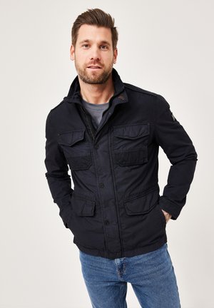 Navy, lightweight jacket with a zip and button closure, four front pockets, and a stand collar. Worn over a gray shirt and blue jeans.
