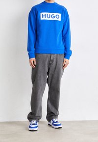 Niebieska bluza z białym logo "HUGO", szare luźne dżinsy oraz niebiesko-białe trampki. Gładka tkanina z ściągaczami na mankietach i u dołu.