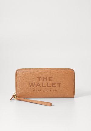 Portafoglio in pelle tan con superficie texture, caratterizzato da scritte in rilievo "THE WALLET" e "MARC JACOBS", e un cinturino da polso. Chiusura con zip.