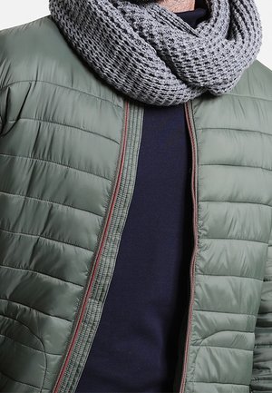 Homme portant une écharpe grise en tricot, un pull bleu marine et une veste matelassée verte non zippée avec une bordure rouge.