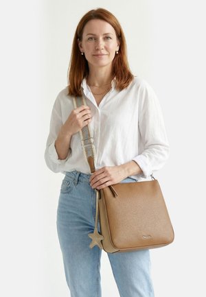 Frau trägt weißes Hemd und blaue Jeans, hält eine braune Ledertasche mit sternförmigem Anhänger vor weißem Hintergrund.