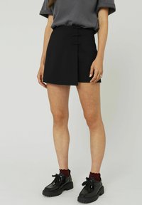 Mini-jupe noire avec un design enroulé, ornée de deux boutons décoratifs. Confectionnée en tissu lisse avec une finition soignée. Associée à des chaussures à plateforme noires.