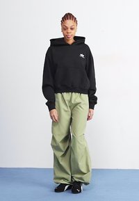 Svart hettegenser med frontlomme, grønne vide bukser laget av glatt stoff, og svarte joggesko. Enkelt design, ingen mønstre.