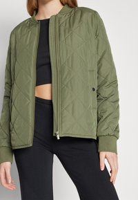 Olivgrön vadderad bomberjacka med ribbade ärmslut, blixtlås och rundad halsringning. Har en slät, matt yta.