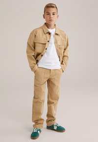 Fant je obrnjen proti spredaj s rokama v žepih rjavih hlač, oblečen v rjav zapenjalni jopič čez belo srajco in z zelenimi supergami z belimi vezalkami.