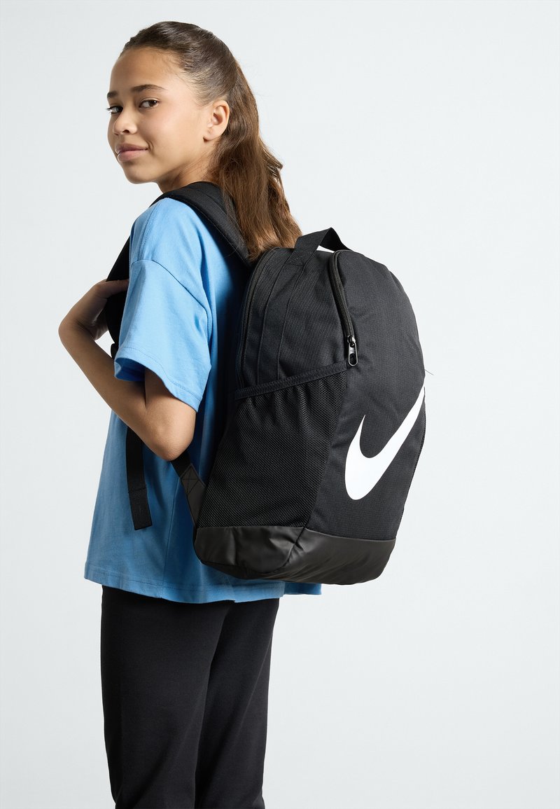 Svart Nike-ryggsäck med en texturerad tyg och en blank svart botten. Har en stor vit logotyp och justerbara remmar.