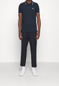 Marinblå polo med vit krage och ärmdetaljer, kombinerad med mörka byxor och vita sneakers. Innehåller en logga på bröstet.