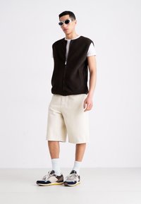 Chaleco negro con cremallera, camiseta blanca, pantalones cortos color crema y zapatillas multicolores. Gafas de sol de estilo vintage completan el atuendo. Diseño minimalista.