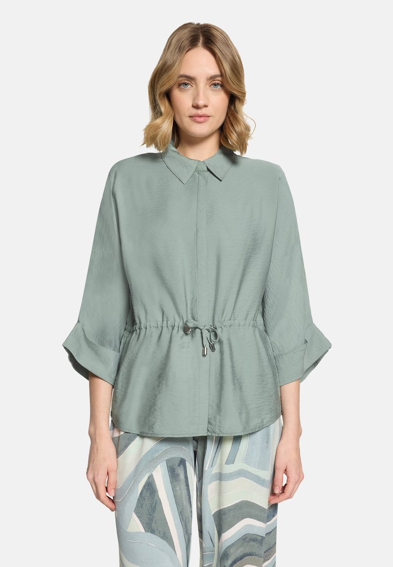 Femme portant un chemisier vert clair à manches longues avec taille à cordon et poignets retroussés, associé à un pantalon large à motifs dans des tons bleus.