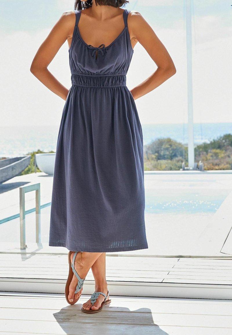 Femme debout à l'intérieur près d'une porte vitrée, portant une robe midi sans manches bleu marine et des sandales tressées bleu clair, jambes croisées.