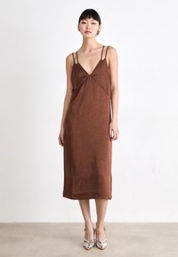 Rochie lungime medie, de culoare maro, cu bretele subțiri, decolteu în V și material texturat. Asortată cu pantofi cu toc înalt, de culoare argintie, evidențiind un design elegant.