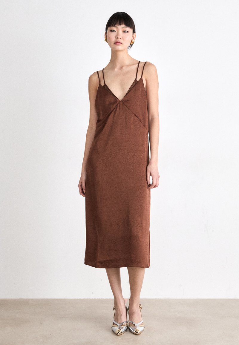Rochie lungime medie, de culoare maro, cu bretele subțiri, decolteu în V și material texturat. Asortată cu pantofi cu toc înalt, de culoare argintie, evidențiind un design elegant.