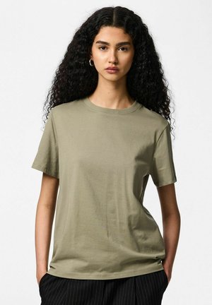 Camiseta de algodón color verde oliva con mangas cortas y cuello redondo, con un ajuste relajado. La persona lleva la mano en el bolsillo.