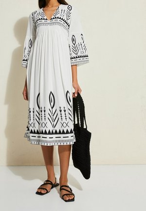 Femme portant une robe midi blanche avec des motifs géométriques noirs, des sandales noires à lanières et tenant un sac cabas tressé noir.