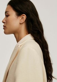 Abrigo de lana beige con cuello de chal, que presenta un detalle de botón único. Pelo largo y ondulado de color oscuro y un pequeño pendiente de aro visible.