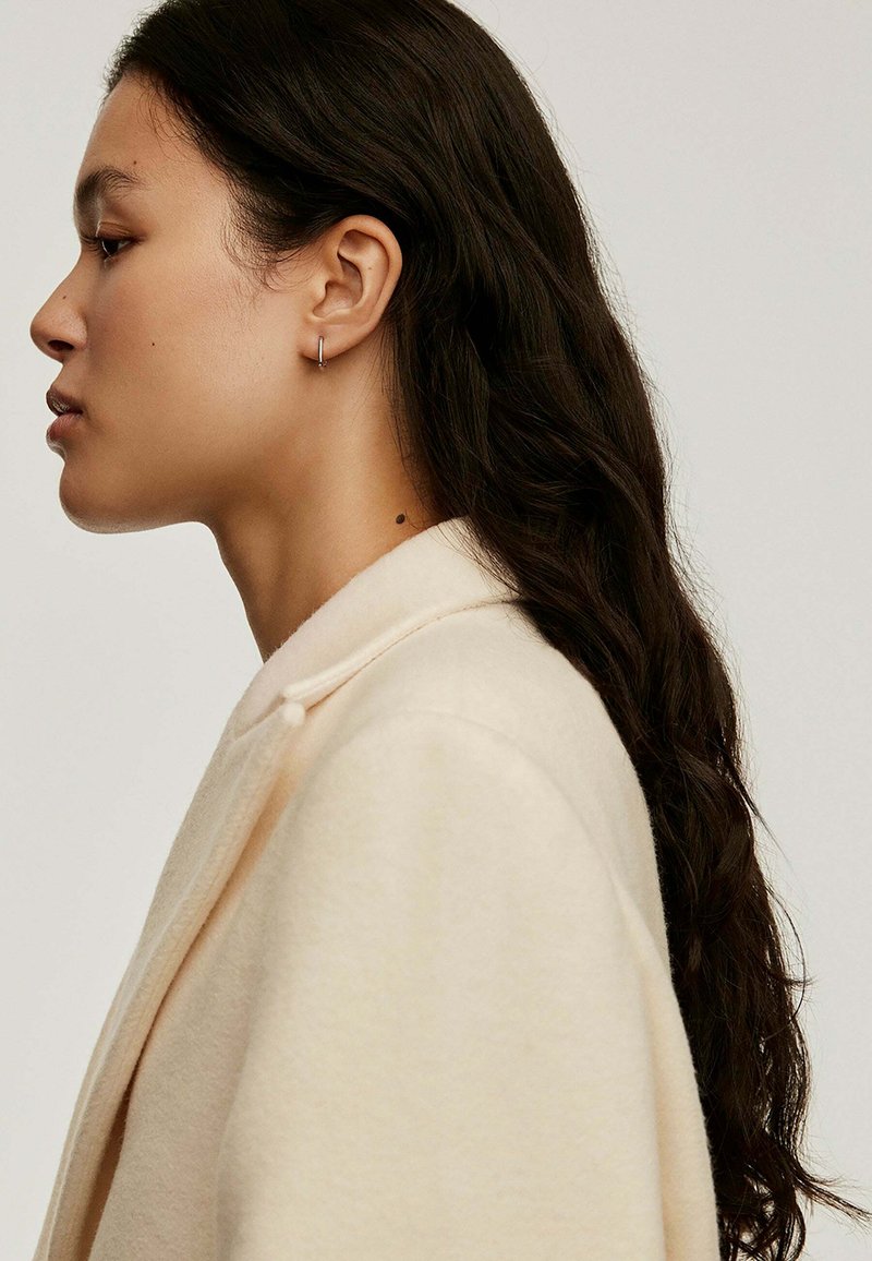 Abrigo de lana beige con cuello de chal, que presenta un detalle de botón único. Pelo largo y ondulado de color oscuro y un pequeño pendiente de aro visible.