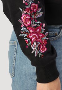 Μαύρη φούτερ με πολύχρωμη κεντημένη λουλουδάτη λεπτομέρεια σε ροζ και κόκκινες αποχρώσεις στο μανίκι. Διαθέτει ριμπ μανσέτα και απαλή υφή υφάσματος.
