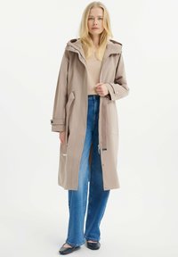 Beige langer Mantel mit Kapuze, vorderen Taschen und verstellbaren Manschetten, getragen über einem hellen Oberteil und blauen Jeans. Glattes, langlebiges Material.