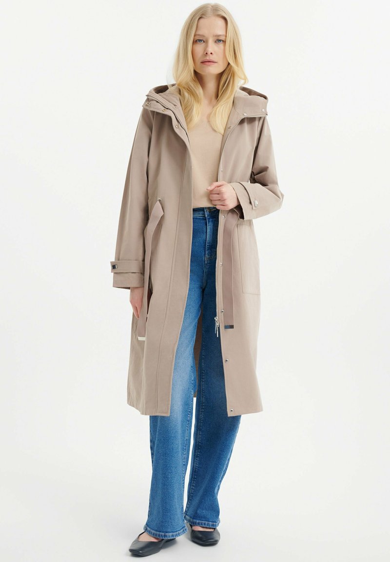 Beige langer Mantel mit Kapuze, vorderen Taschen und verstellbaren Manschetten, getragen über einem hellen Oberteil und blauen Jeans. Glattes, langlebiges Material.