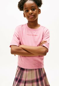Lyserød bomulds T-shirt med rund halsudskæring, prydet med "TOMMY HILFIGER" logoet i midten. Modellen bærer en rutet plisseret nederdel.