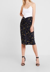 Jupe crayon bleu marine avec un imprimé floral jaune, dotée de détails froncés. Portée avec un débardeur blanc et des escarpins noirs à lanières.