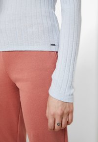 Ljusblå ruggad långärmad topp i kombination med rosa, mjuka byxor. Anmärkningsvärda detaljer inkluderar en liten etikett på toppen av plagget och en tatuering på handen.