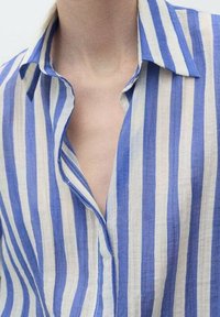 Chemise en lin rayée bleu et blanc, dotée d'un col et d'une patte de boutonnage, avec une coupe décontractée et une texture douce. Les rayures varient en largeur.