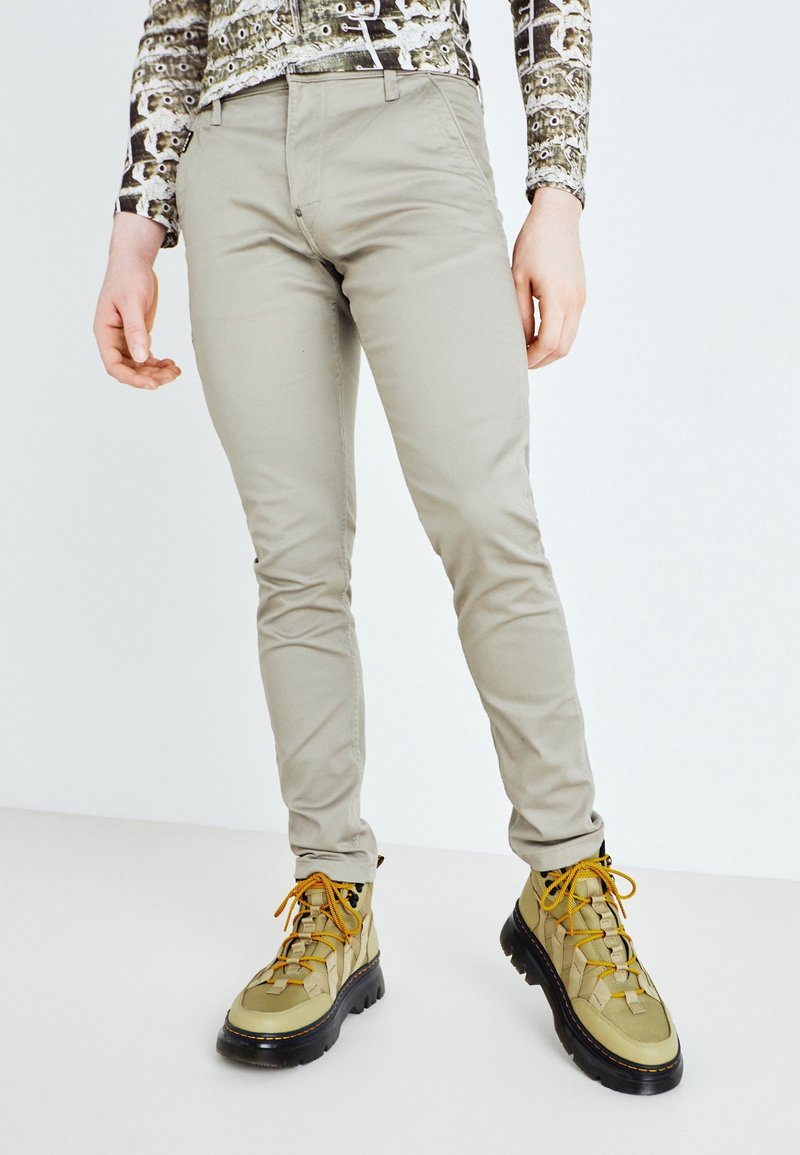 Personne portant un pantalon beige slim et des chaussures de randonnée vert clair avec des semelles épaisses noires et des lacets jaunes.
