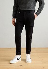 Černé nákladní kalhoty z elastické tkaniny, s bočními kapsami, zúženým střihem, kombinované s bílými kotníkovými teniskami.