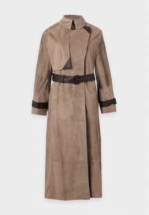 Frau trägt einen langen taupefarbenen Trenchcoat mit schwarzen Lederakzenten und Gürtel, kombiniert mit weiten dunklen Hosen und schwarzen Schuhen, stehend vor weißem Hintergrund.