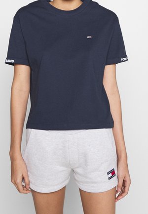 Osoba nosząca granatowy krótki t-shirt Tommy Jeans oraz jasnoszare szorty z logo Tommy Jeans przy brzegu, stojąca na jednolitym tle.