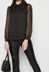 Svart blus med genomskinliga ärmar, knappstängning framtill och en pärlkrage. Matchad med högblanka svarta leggings och en liten svart handväska.