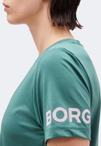 Προφίλ ενός ατόμου που φοράει μια βεραμάν κοντομάνικη μπλούζα με τη λέξη "BORG" τυπωμένη με μεγάλα ροζ γράμματα στο μανίκι.