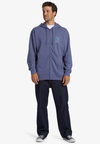 Sudadera azul con cremallera y bolsillo frontal, con cordones. Cuenta con un logotipo gráfico en el lado izquierdo del pecho. Combinada con jeans oscuros y zapatillas.