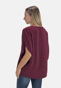 Top color amaranto con maniche a pipistrello, tessuto strutturato, scollo ampio e spacchi laterali. Abbinato a jeans blu, mostrato da dietro.