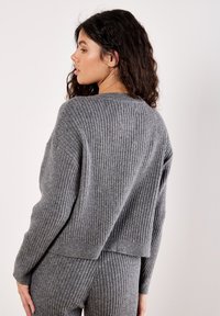 Frau mit langen lockigen Haaren, die einen grauen gerippten Strickpullover und passende gerippte Strickhosen trägt, steht seitlich vor einem schlichten Hintergrund.