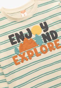Bawełniany T-shirt z kremowym tłem, turkusowymi paskami i kolorowymi aplikacjami z tkaniny. Zawiera napisy "CIESZ SIĘ" oraz "BADANIE" z wizerunkami słońca, gór i drzew.