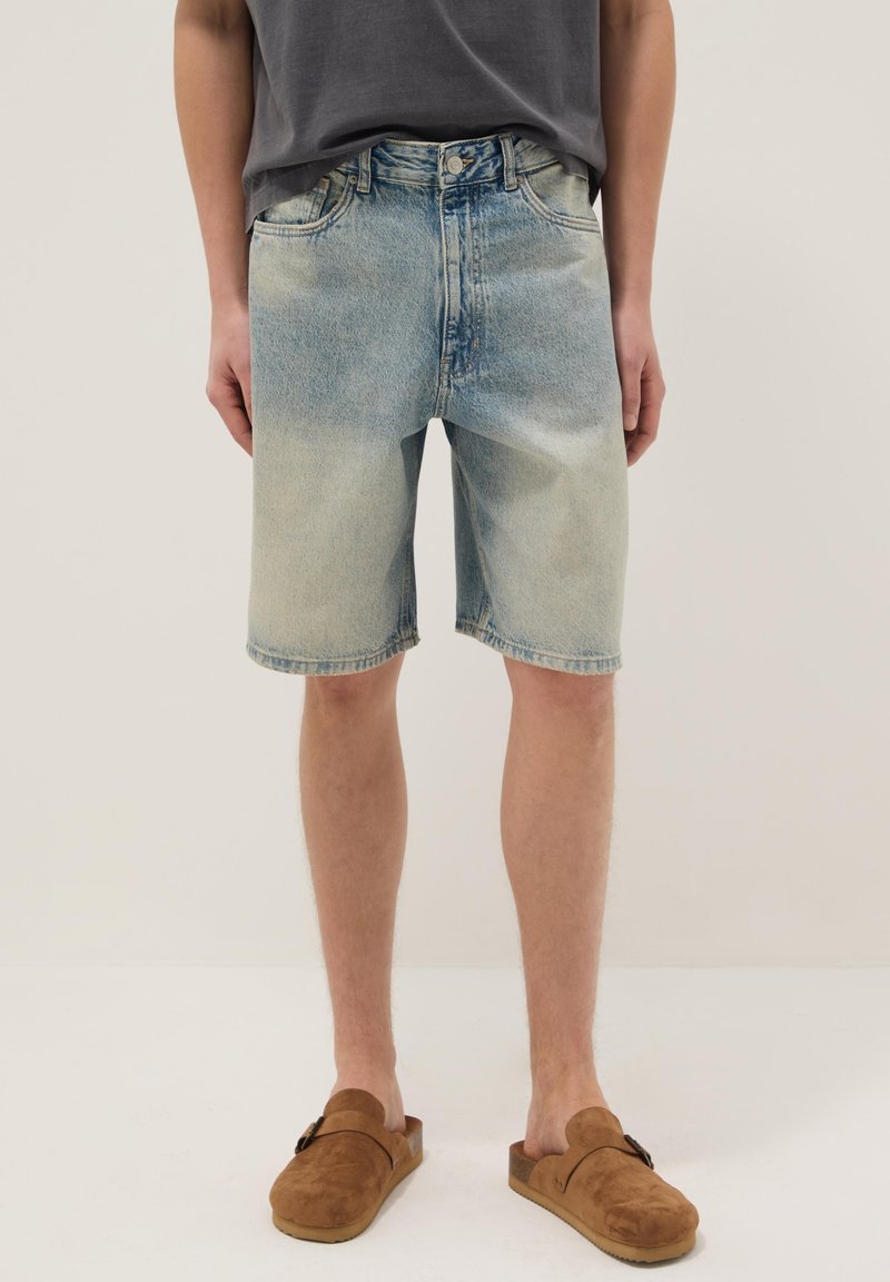 Hombre con pantalones cortos de mezclilla azul claro, camiseta gris oscuro y zapatos deslizantes de ante marrón, de pie contra un fondo liso.