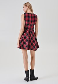 Abito senza maniche a scacchi rosso e nero con un corsetto aderente e una gonna svasata; cucitura posteriore visibile e chiusura con bottoni; abbinato a stivali neri alla caviglia.