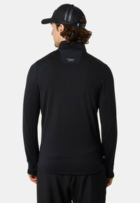 Zwarte longsleeve performance top met hoge kraag, gestructureerde stof en logo op de rug. Heeft een getailleerde pasvorm en zoom.