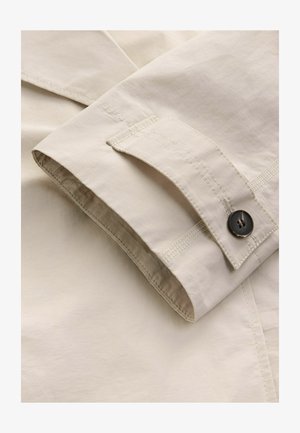 Helles beiges Gewebe mit einer glatten Textur. Verfügt über einen gefalteten Ärmelabschluss mit einem Knopf und kontrastierenden Nähten, die das Design hervorheben.