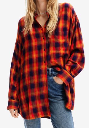 Femme portant une chemise à carreaux oversize rouge, orange et bleu marine avec poche avant, associée à un jean bleu, main dans la poche, visage non visible.